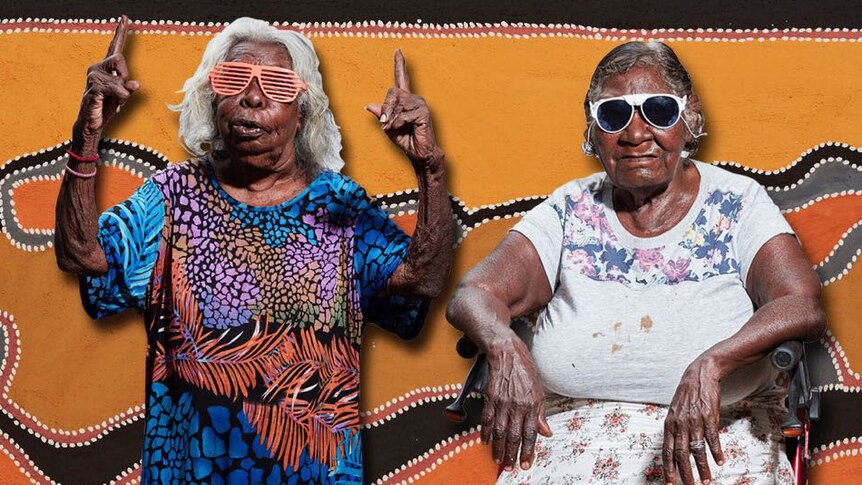 Two Aboriginal women wearing sunglasses, one seated and one with raised hands,  in front of a dot painting.