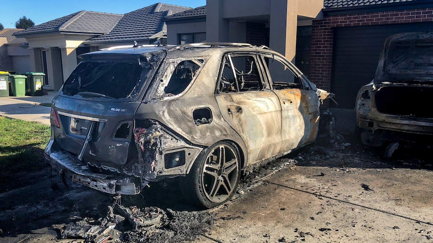 Two significantly burnt out and damaged cars in a driveway, with suburban homes in the background.