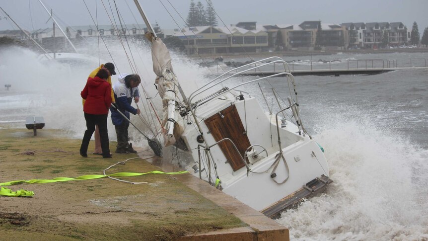 Three people try to tie up a small yacht after it washed into a harbour wall at a sailing club in Bunbury.