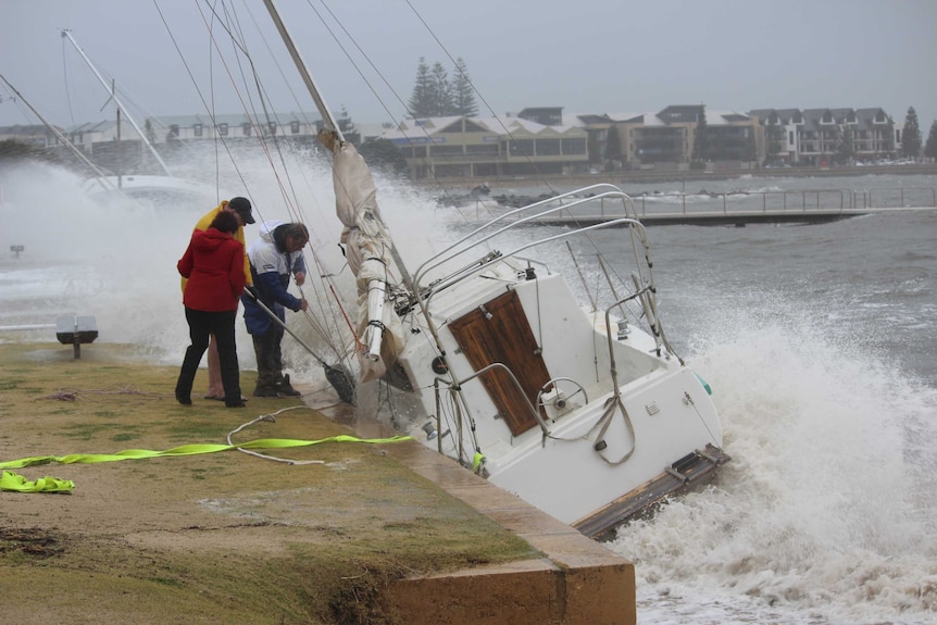 Three people try to tie up a small yacht after it washed into a harbour wall at a sailing club in Bunbury.