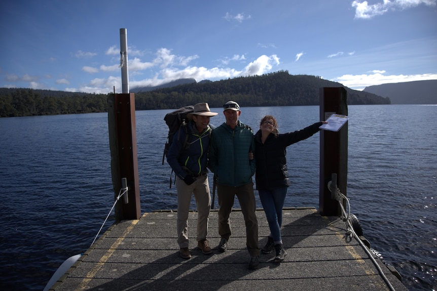 Two men and a woman standing at the end of a pier, smiling, with a lake and hills in the background. 