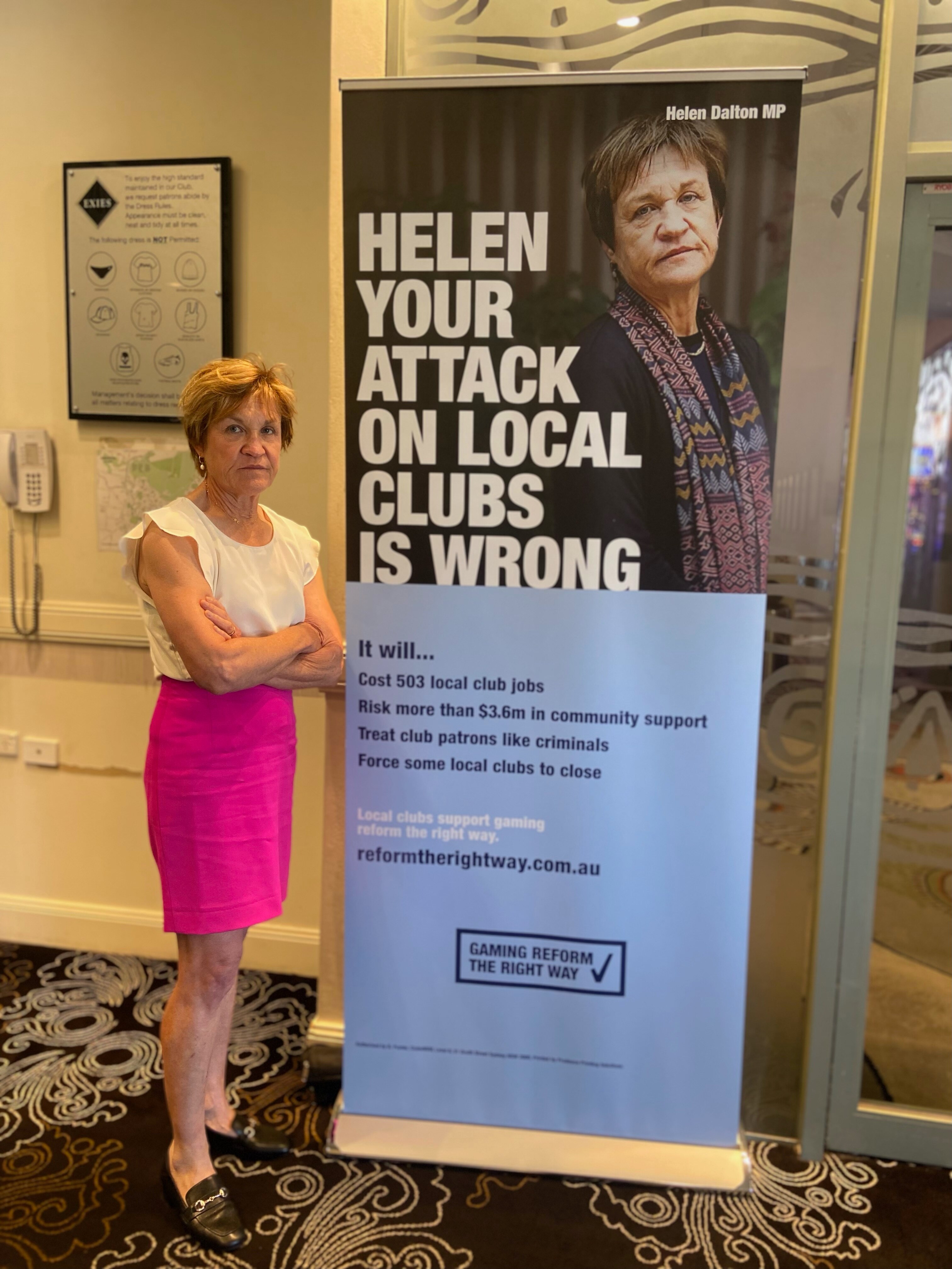 A woman with arms crossed stands next to a sign that has her photo and says 'Helen your attack on local clubs is wrong'