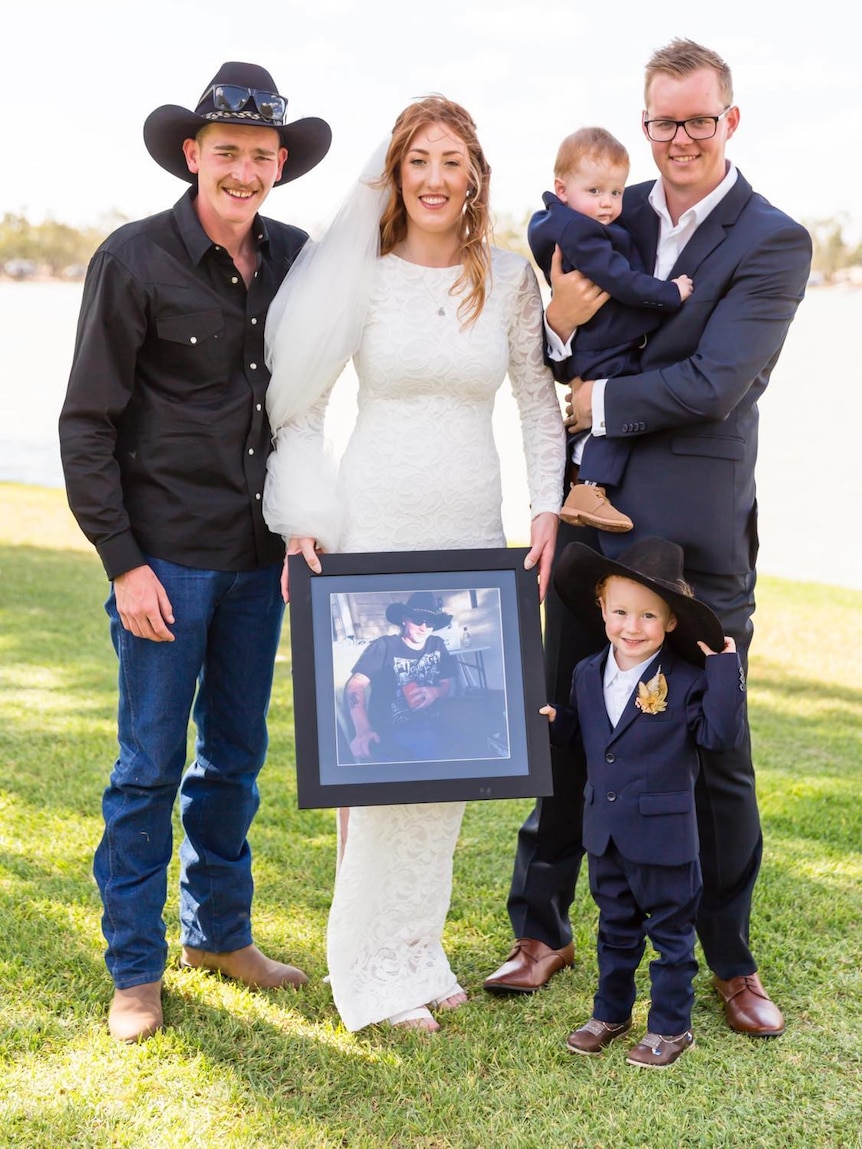 A bride holds a picture of a man by a lake, standing with two men in shirts and a toddler in a hat.