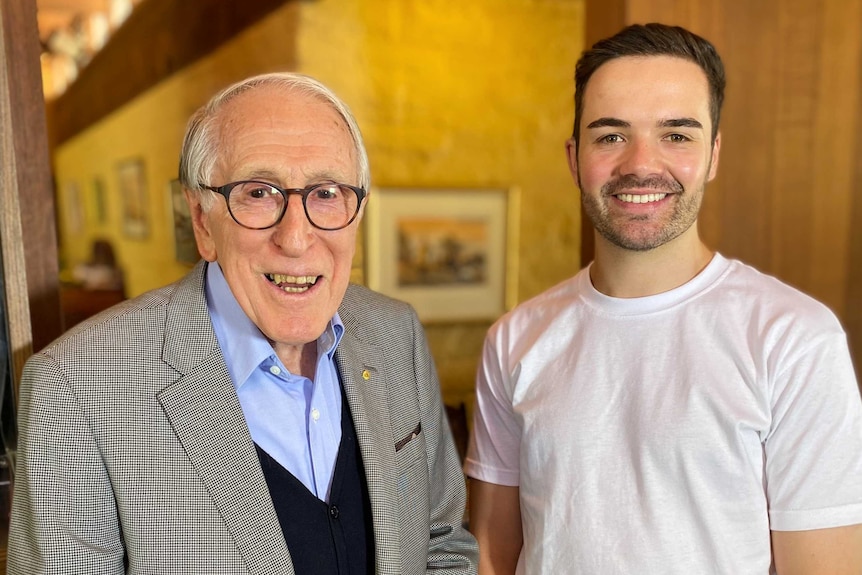 An elderly man with glasses, wearing a suit jacket stands next to a younger man in a white t-shirt.