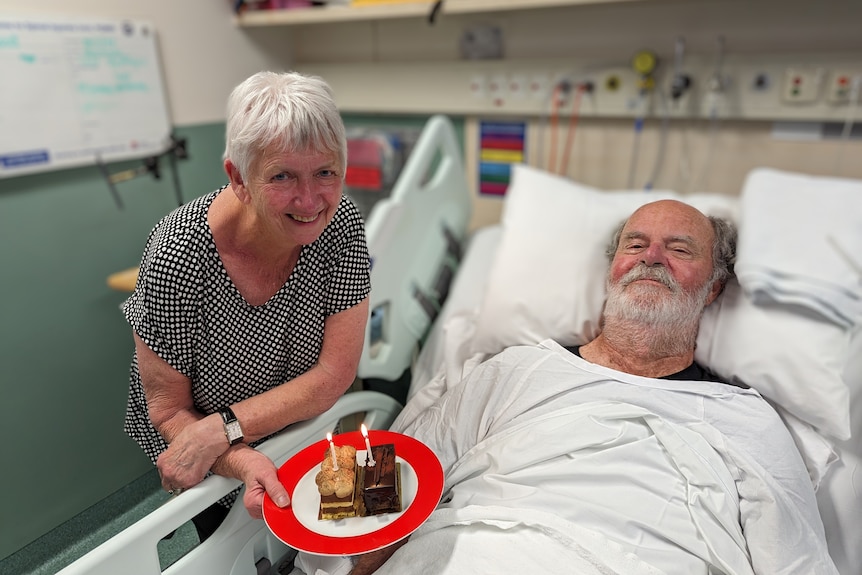 Man with white beard lying in hospital bed with a woman beside him holding cake. 