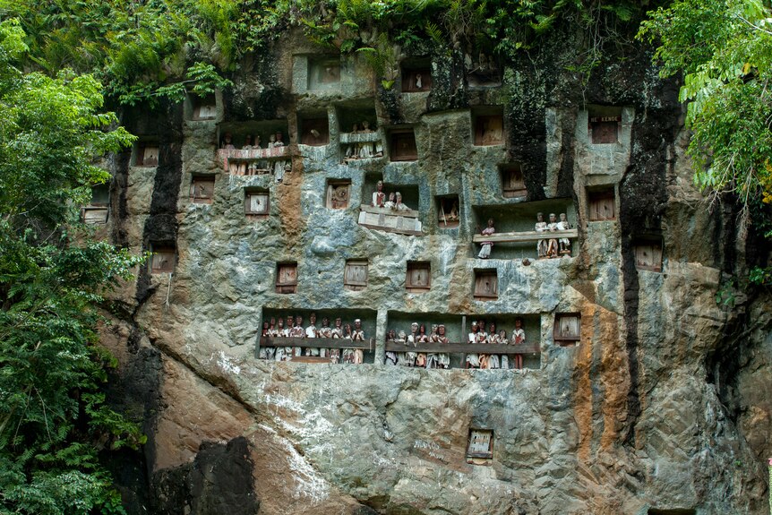 A cliff face with balconies features carved wooden figures standing in rows. 