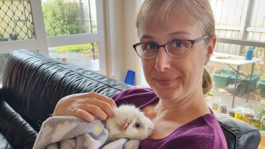 A blonde woman in her 30s wearing glasses holds a white guinea pig