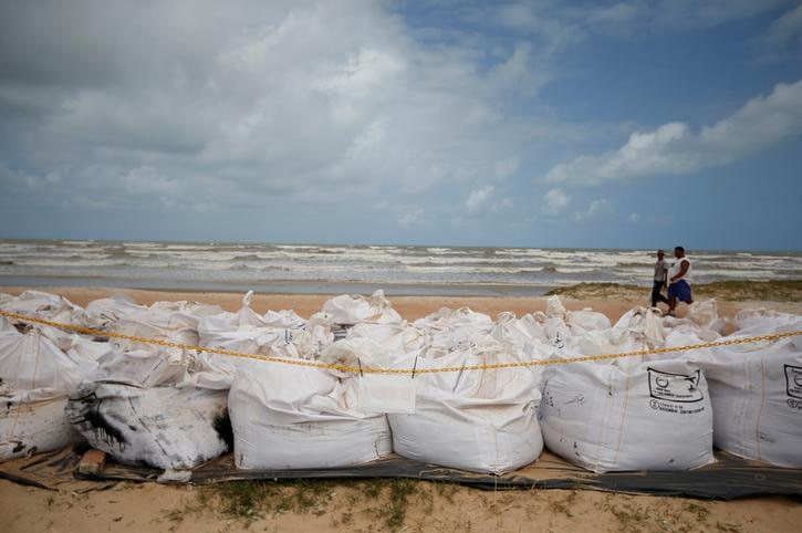 Tens of bags are seen filled with the beach in the background. Two people walk on the sand between the bags and the sea.