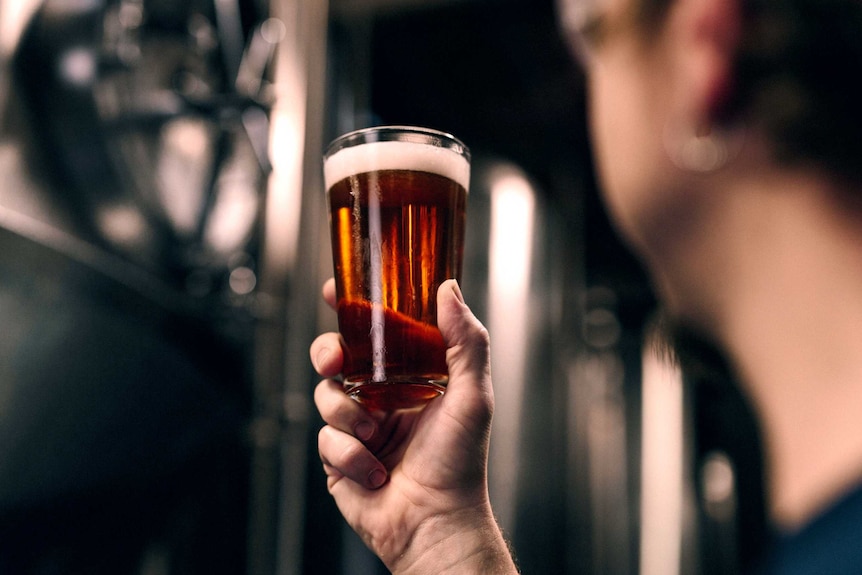 A man holds up a glass of beer against a backdrop of metal brewing vats.