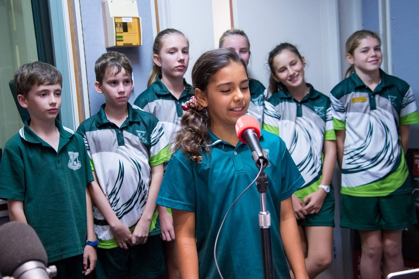 School children wait in line while a girl speaks into a microphone.