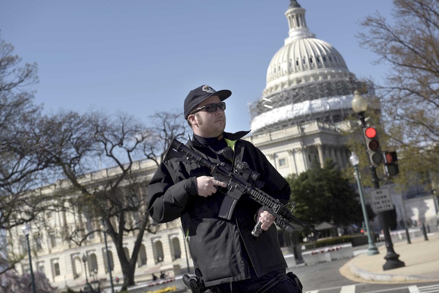 A police officer holding a large rifle stands in front of the US Capitol building.