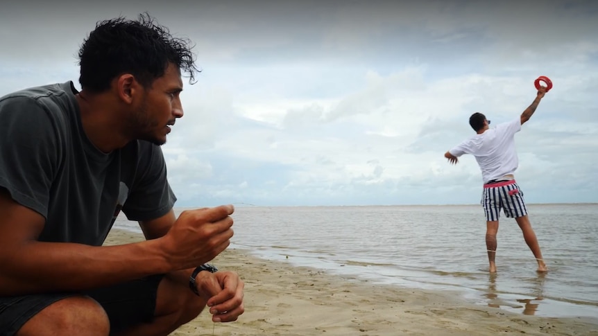 One man squatting on the sand and looking out at the ocean while another man stands in the shallows fishing