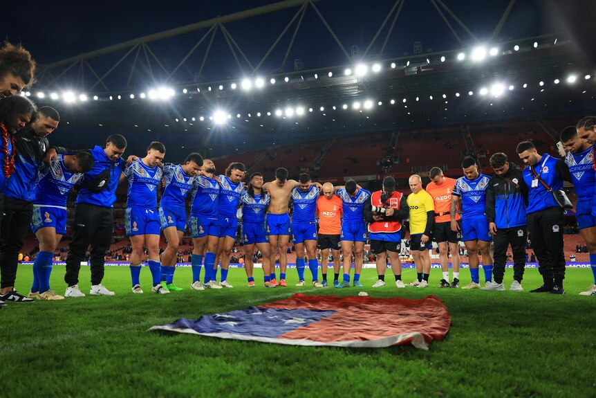 The Samoan rugby league team stand with heads down and eyes closed as they gather around the Samoan flag after a big win.