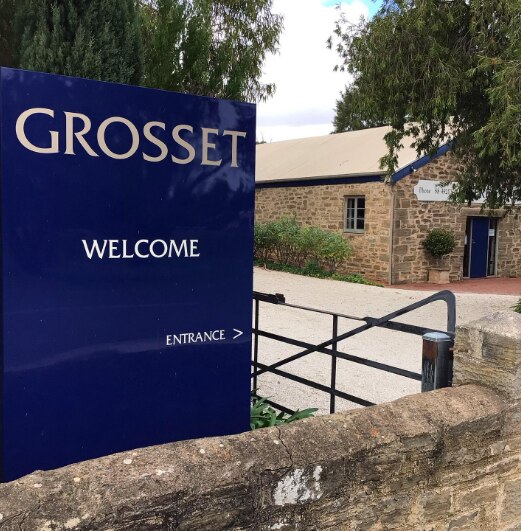 A blue sign with Grosset, welcome, entrance in front of a gate and an old brick building, greenery in front and side, blue door.
