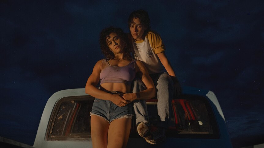 A film still of Katy O'Brian and Kristen Stewart, together in the cab of a blue truck, wearing 80s-style clothing.