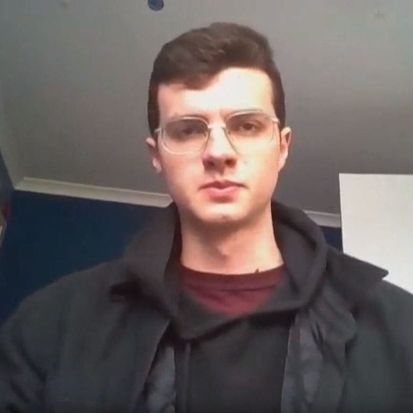 A man stares down the barrel of a camera while sitting in a room, wearing all black.