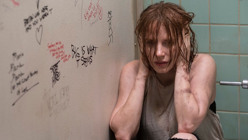 A distraught woman with wet hair and tattered clothes crouches in graffitied toilet stall while covering both ears with hands.