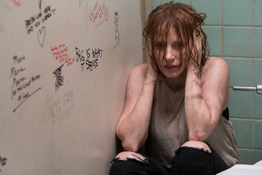 A distraught woman with wet hair and tattered clothes crouches in graffitied toilet stall while covering both ears with hands.