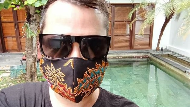 A man with brown hair wearing black sunglasses and a black mask with a colourful flower pattern stand in front of a pool.
