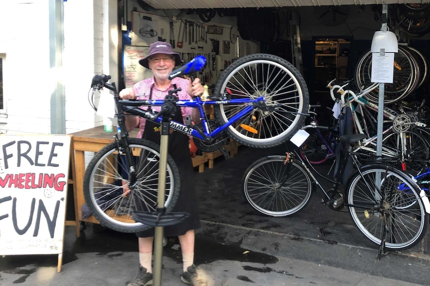 A smiling man in an apron holds up a bicycle in front of a garage with a sign out front that says 'Free Wheeling Fun'.