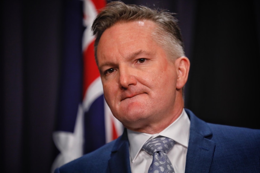 A close shot of Chris Bowen's face, looking serious, an Australian flag and blue curtain on the wall behind him.