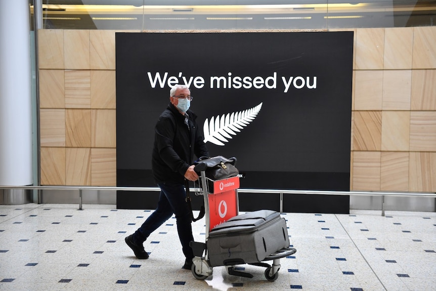 A man in a mask pushes a suitcase trolley at Sydney airport, in front of a sign with silver fern that says: "We've miss you".