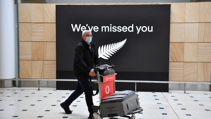 A man in a mask pushes a suitcase trolley at Sydney airport, in front of a sign with silver fern that says: "We've miss you".