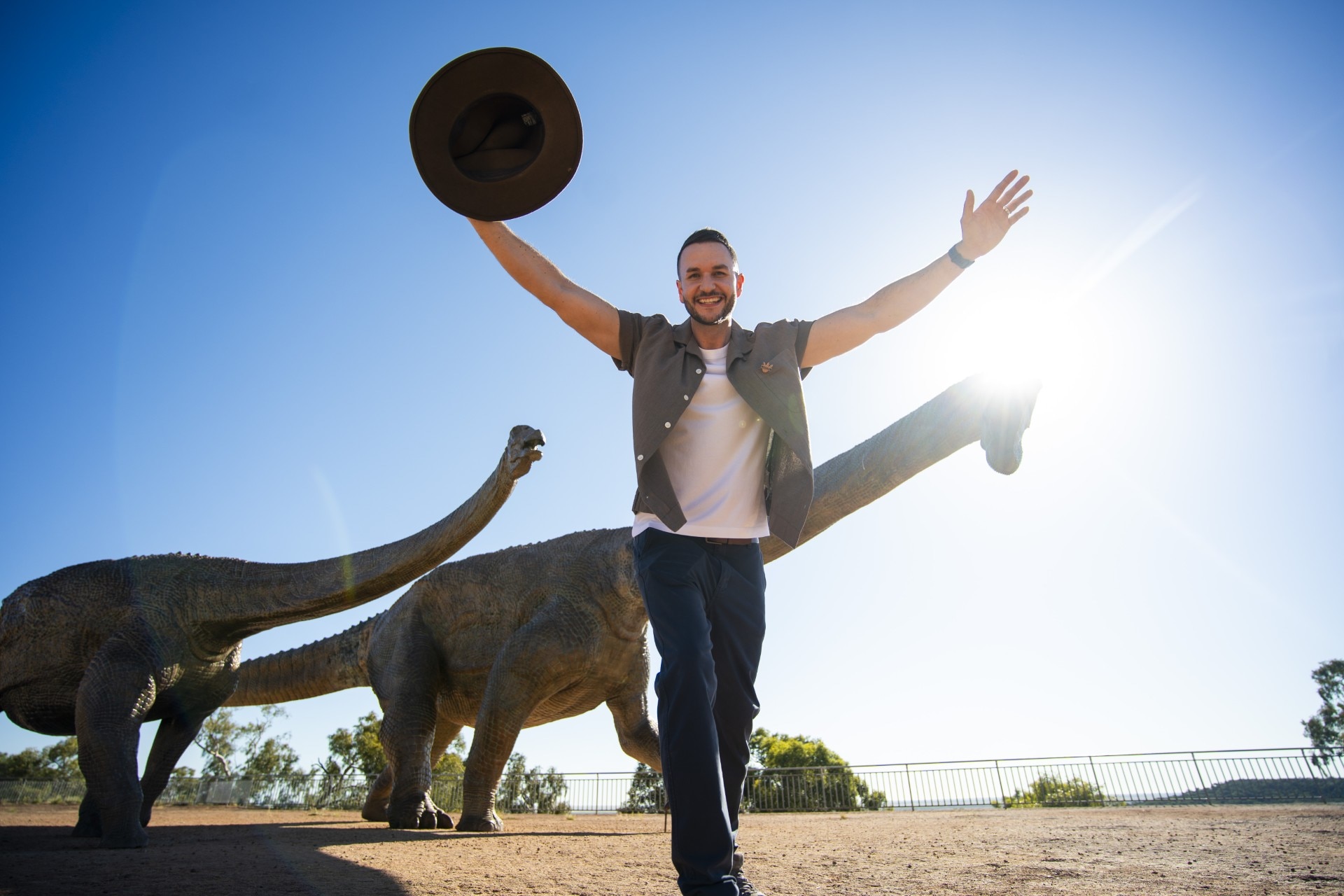 Um homem sorridente e de cabelos escuros segura o chapéu no ar enquanto fica perto de algumas estátuas de dinossauros em uma área deserta.