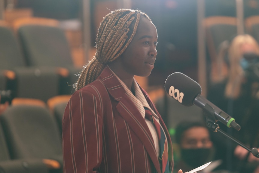 A female student with plaited hair and a burgundy striped blazer stands at a microphone 