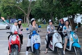 Six young women smile as they sit on motor scooters in front of a park.