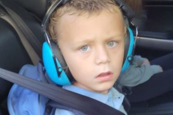 A tight mid shot of a young boy wearing ear muffs and a blue shirt sitting in a car.
