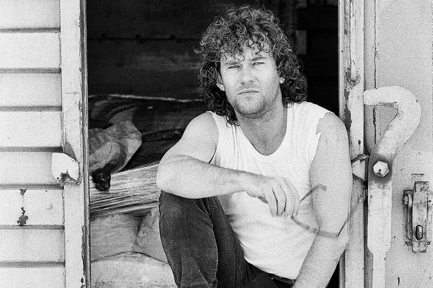 Jimmy Barnes sits with his elbow on his knee in the door of an old sugar cane wagon.