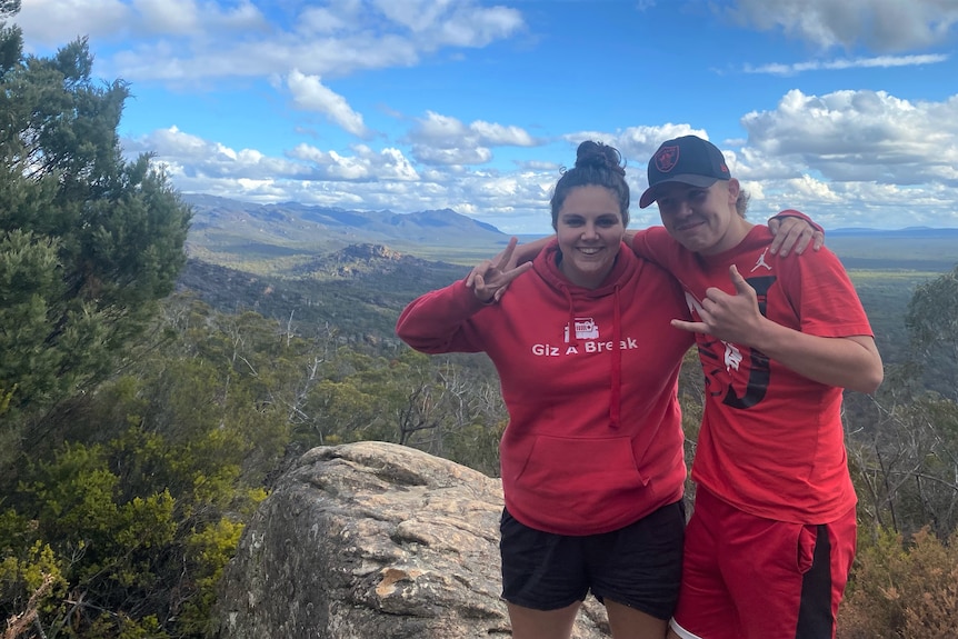 A woman in a red hoodie and a boy in a red singlet stand with their arms around one another in front of a view of mountains.