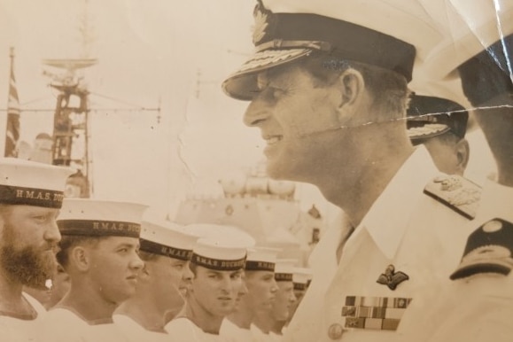A vintage photo of a sailor standing in a line being address by a senior officer, with one person looking at the camera
