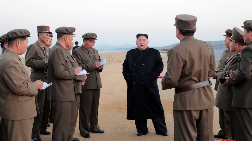 Kim Jong-un stands on a dirt road surrounded by military personnel taking notes.