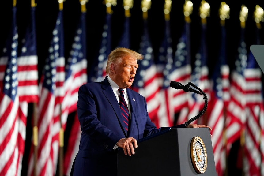 A man in a suit stands at a lectern with US flags behind him.