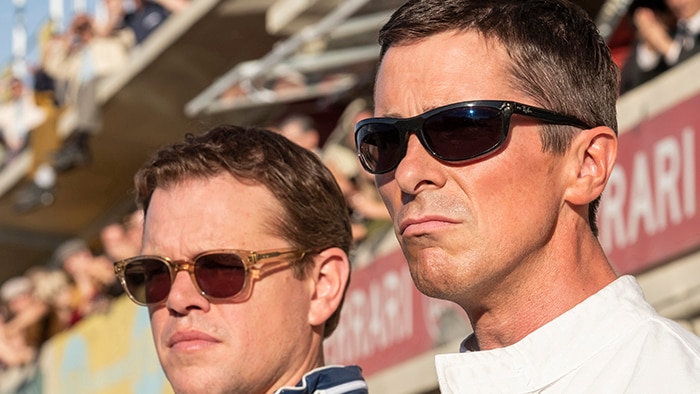 Matt Damon and Christian Bale wearing sunglasses stand side by side in front of racing track spectator stands.