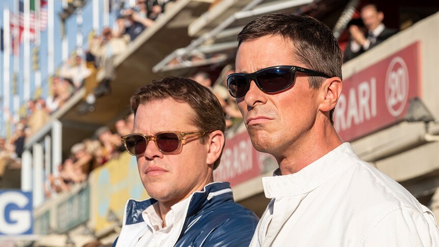 Matt Damon and Christian Bale wearing sunglasses stand side by side in front of racing track spectator stands.