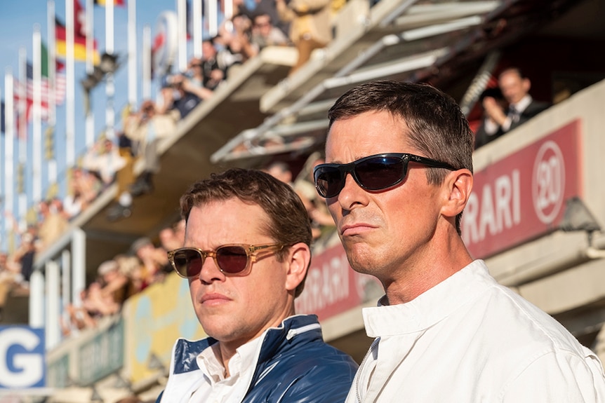 Matt Damon and Christian Bale wearing sunglasses stand side by side in front of racing track spectator stands.