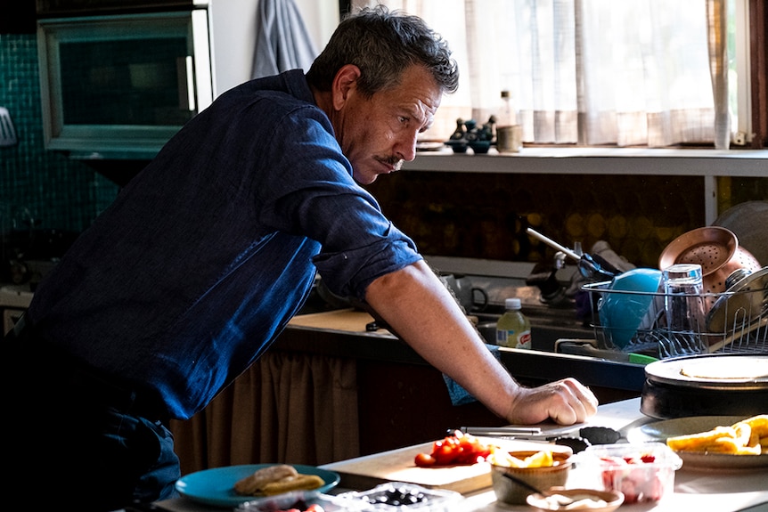 A man with greying hair and moustache stands in denim shirt leans forward on kitchen bench in mid-prep near window on sunny day.
