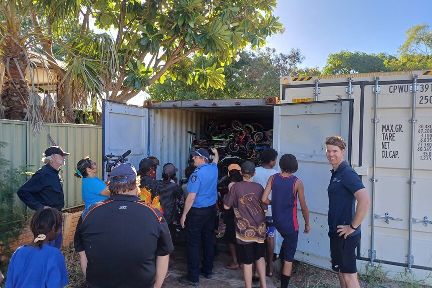 police, children and government workers stand outside a shipping container full of bikes