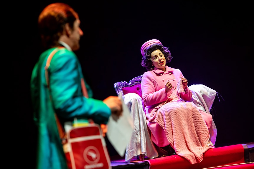 Monique Sallé onstage as Queen Elizabeth, dressed in all-pink with matching toque hat, knitting and singing from a chair.