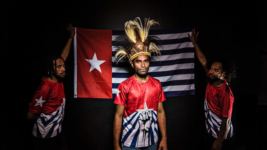 Colour photograph of activist Ronnie Kareni standing in front of the Morning Star flag held up by Yoshua and Sam Roem.