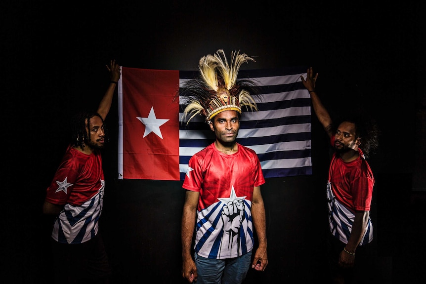 Colour photograph of activist Ronnie Kareni standing in front of the Morning Star flag held up by Yoshua and Sam Roem.