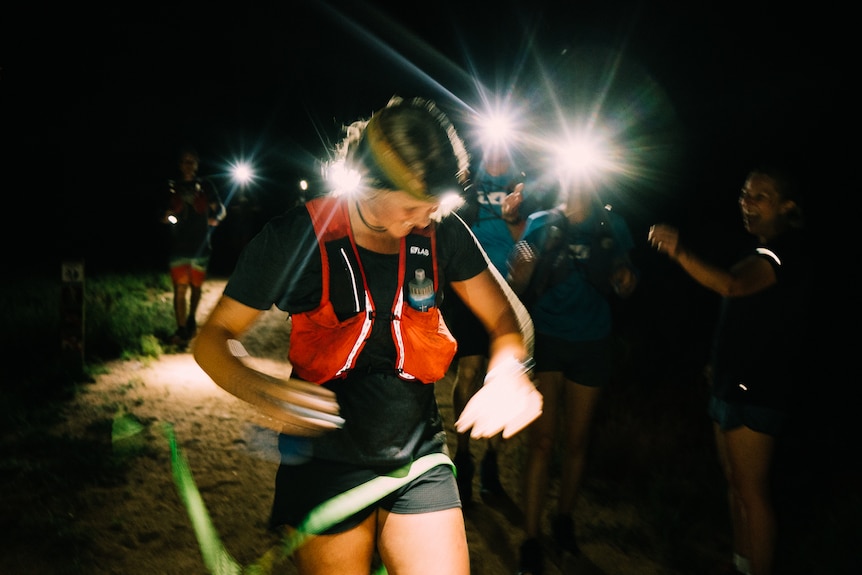 night time image of blond haired woman wearing headtorch running wearing red running vest. Runners with headtorches behind her. 