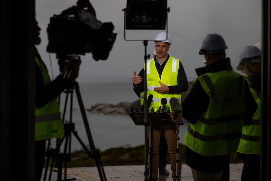 A man wearing a high vis vest and hard hat stands in front of media microphones and lights, with camera operators in shadow