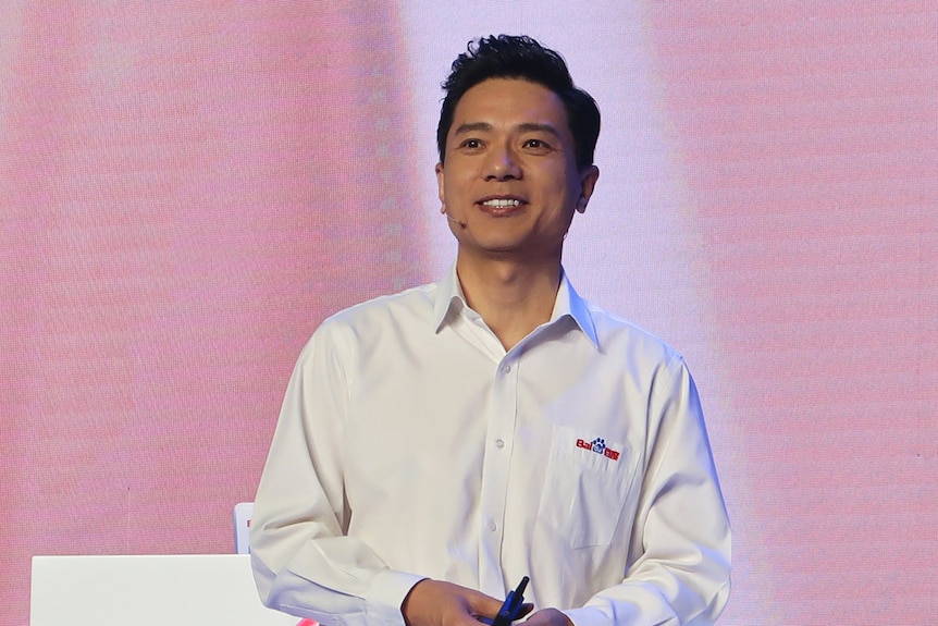 A young man wearing a white collared shirt stands on stage in front of a pink background with hands clasped