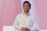 A young man wearing a white collared shirt stands on stage in front of a pink background with hands clasped