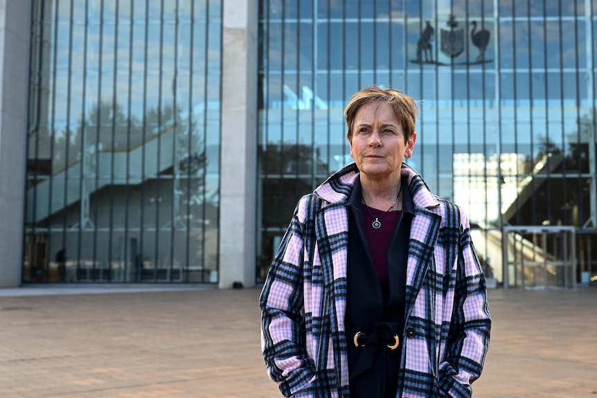 Robyn Milera, with short brown hair, check coat and black shirt, stands with serious expression in front of large building.