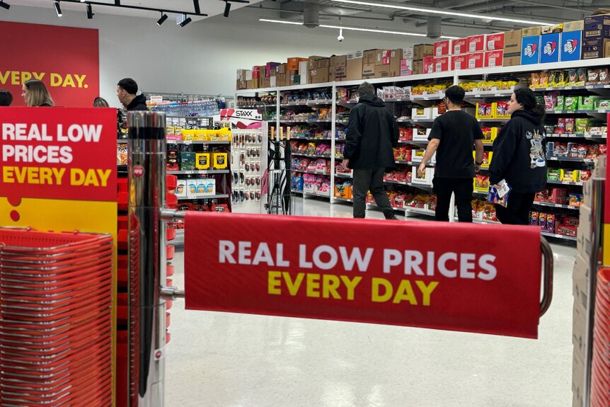 A red store entry barrier that says "real low prices every day" with people queuing near shelves stocked with snacks.